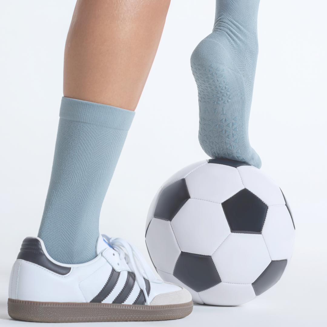 Person wearing light blue socks and white sneakers with black stripes, standing next to a soccer ball on a light gray background.