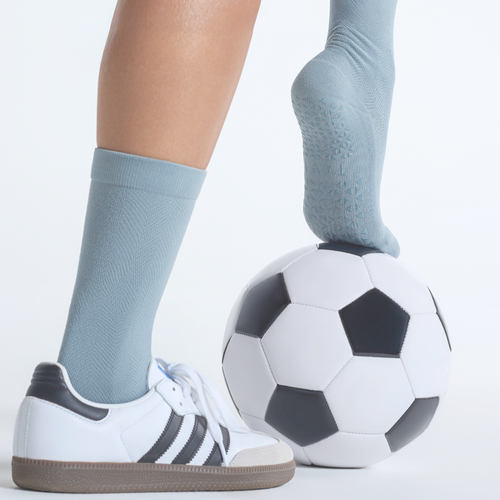 Person wearing light blue socks and white sneakers with black stripes, standing next to a soccer ball on a light gray background.